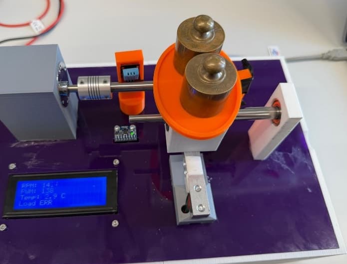 The finished rotary test rig on a purple acrylic base: grey motor housing, silver coupling, orange gearbox with two brass weights on its load plate, white end bearing, and a blue LCD showing live readings.
